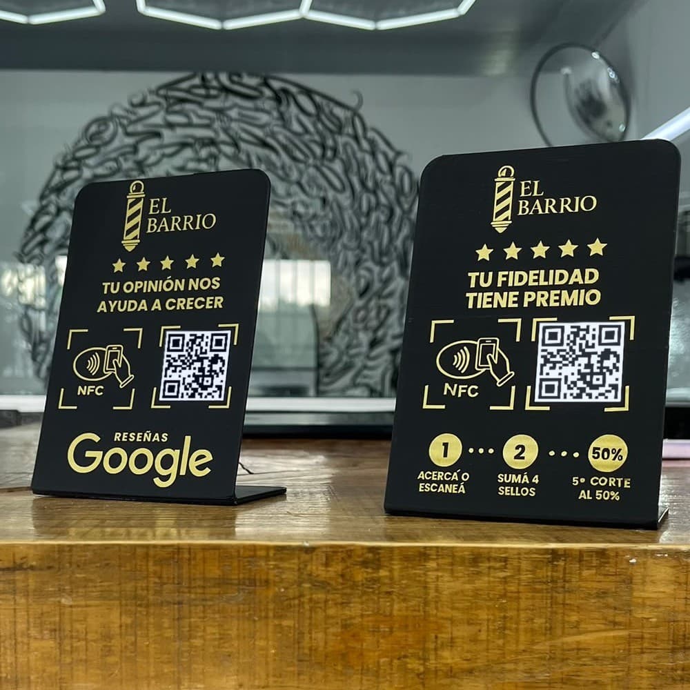 Dos stands negros con letras doradas de El Barrio Barbershop sobre el mostrador: uno para dejar la reseña en Google y otro para la tarjeta de sellos, los dos con NFC y código QR