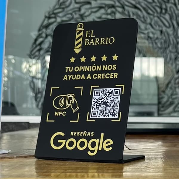 Stand negro con letras doradas de El Barrio sobre el mostrador de la barbería: «Tu opinión nos ayuda a crecer», con NFC, código QR y el logo de reseñas de Google