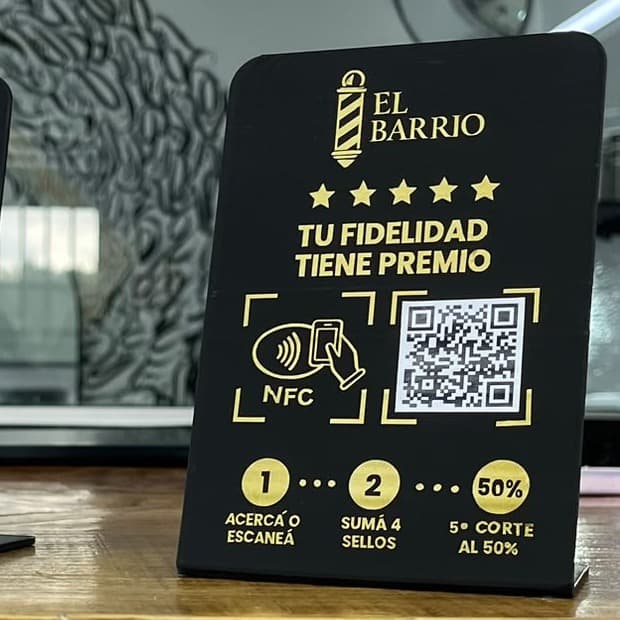 Stand negro con letras doradas de El Barrio sobre el mostrador de la barbería: «Tu fidelidad tiene premio», con NFC, código QR y los pasos acercá o escaneá, sumá 4 sellos, 5.º corte al 50 %