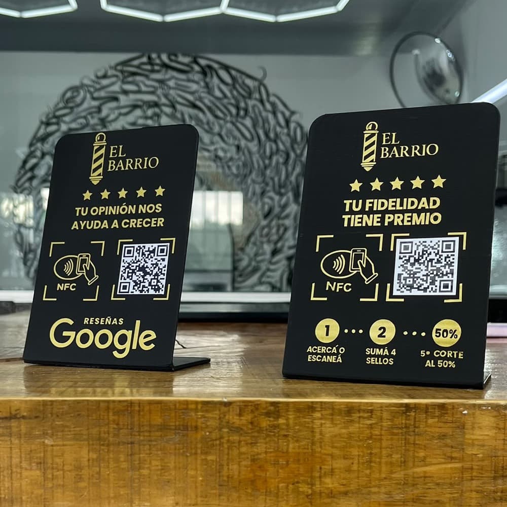 Dos stands negros con letras doradas de El Barrio Barbershop sobre el mostrador: uno para dejar la reseña en Google y otro para la tarjeta de sellos, los dos con NFC y código QR